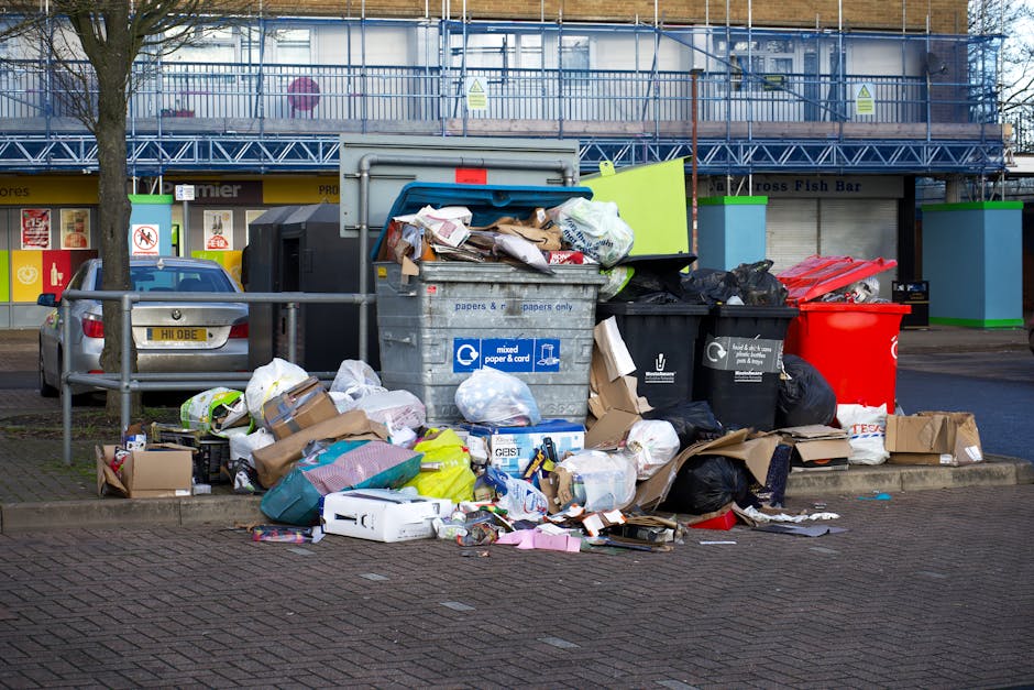 A collection of overflowing waste and recycling bins located on a paved pavement area in Poplar, with various types of rubbish including cardboard boxes, plastic bags, black bin bags, paper, and packaging materials surrounding the bins. The waste includes flattened boxes, loose papers, and plastic containers, some of which are spilling onto the ground. Behind the bins, there is a building under construction with scaffolding and protective netting, along with a row of shopfronts visible at street level, including a fish bar. A grey car is parked nearby behind a metal railing, which separates the waste area from the street. The scene is lit with natural daylight, and the rubbish appears prepared for collection or disposal, reflecting typical household or commercial waste accumulated during a home relocation or decluttering process, relevant to the services of Man with Van Poplar in scheduling council or private waste removal.