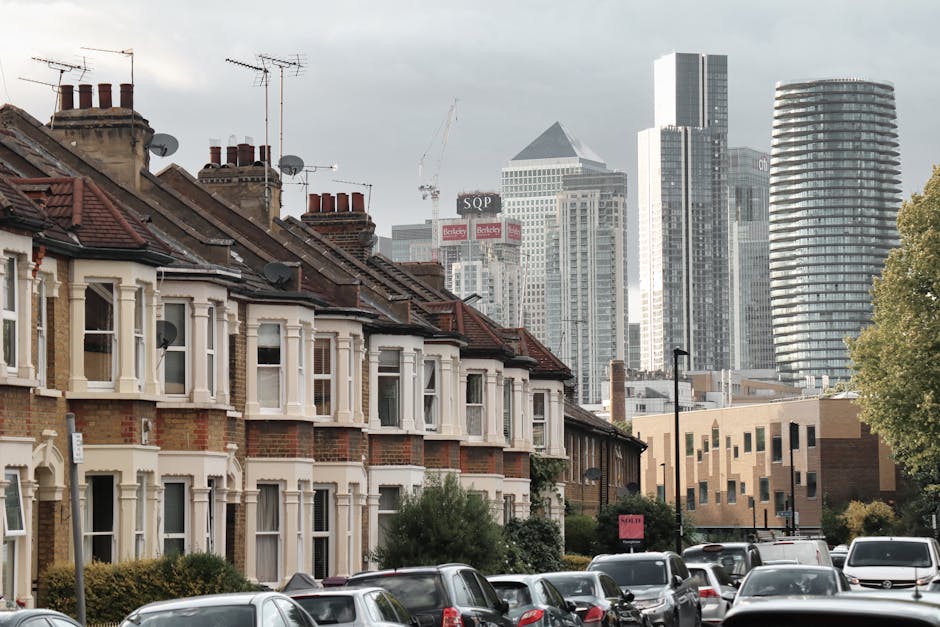 A row of traditional two-storey terraced houses with brick facades and chimney stacks, featuring white bay windows and red tiled roofs, are visible in the foreground. Several satellite dishes are mounted on the rooftops, and the houses are closely aligned along a residential street with parked cars lining the pavement. In the background, a modern city skyline with tall skyscrapers, including a glass high-rise and a uniquely curved building, stands against a cloudy sky. The scene depicts an urban environment combining residential housing with nearby commercial and office buildings, typical of a home relocation setting where furniture transport and packing might take place, supported by professional removals services such as those offered by Man with Van Poplar.