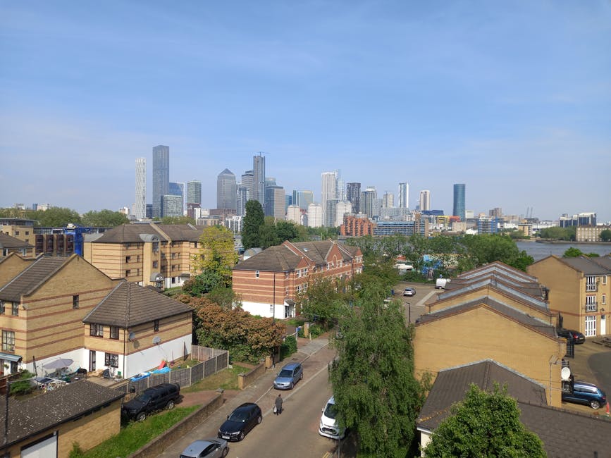 A daytime view over residential houses with brown brick walls, pitched tiled roofs, and small gardens, leading toward a modern city skyline with numerous high-rise office towers and skyscrapers under a partly cloudy blue sky. The foreground features parked cars along a narrow street, some greenery including trees and shrubbery, and a pedestrian walking on the pavement. The image captures an urban environment where home relocation and furniture transport might take place, highlighting the contrast between local housing and the distant business district, supported by the presence of everyday street activities. Man with Van Poplar offers removals and volume logistics services suitable for such moving and packing operations in the area.