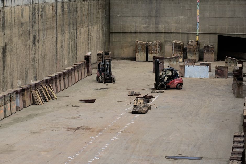 An indoor industrial space used for furniture transport and home relocation preparations, featuring an expansive concrete floor partially marked with white dotted lines indicating loading zones. Two small forklifts are positioned towards the center, one black and one red, amidst stacks of wooden pallets and metal containers. Several wooden pallets and planks are scattered across the area, with some leaning against the walls and others lying flat on the ground. The space is enclosed by high, weathered concrete walls, with yellow, blue, and pink vertical measuring markers attached to the back wall. Bright, natural light illuminates the environment, possibly from openings outside the frame. The arrangement appears to be set up for loading or unloading furniture or large household items, with a focus on efficient move logistics supported by [COMPANY_NAME]’s professional removal services, demonstrating the logistical aspects of furniture transport and packing during home removals.