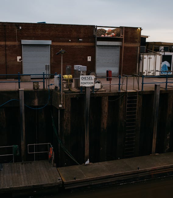 A waterway with a brick industrial building in the background featuring two grey roller shutter doors and a sign reading 'DIESEL PONTOON' mounted on a pole near the edge of the dock. The dock area is guarded by blue metal railings and has a wooden walkway extending over the water. A set of metal stairs provides access from the dock to the water level. The scene is illuminated by ambient outdoor lighting, suggesting early evening or dusk, with no visible vehicles or moving equipment. This setting reflects the loading process for house removals or furniture transport services, as offered by Man with Van Poplar, highlighting the loading area at a property involved in home relocation or packing and moving activities.
