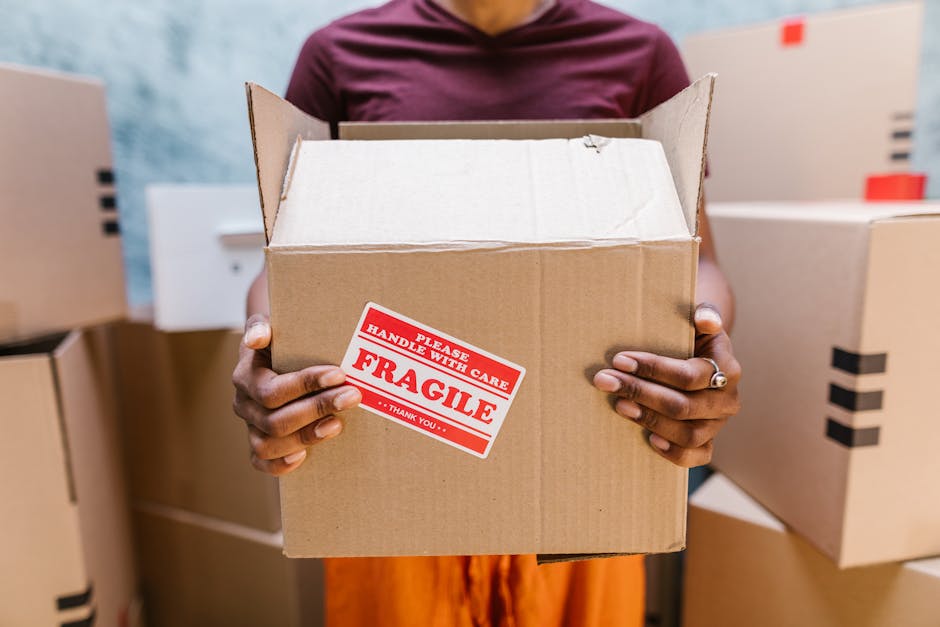 A person with dark skin, wearing a maroon shirt and a ring on their left hand, is holding an open cardboard box labeled 'Fragile' with a red and white warning sticker that reads 'Please Handle With Ca