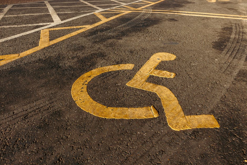 Close-up of a yellow-painted wheelchair accessible parking symbol on a dark asphalt surface in a parking lot. The symbol, showing a stylized image of a person in a wheelchair, is prominently displayed in the foreground. Surrounding the symbol, various parking space markings and lines are visible, including diagonal yellow stripes and white boundary lines. In the background, part of a vehicle access area and an outdoor environment with textured asphalt are visible, indicating an accessible parking zone likely used for customer or client parking related to house removals or relocation services. The scene is lit by natural daylight, providing clear visibility of the textured paving and markings, consistent with parking areas associated with property access points used during home relocations or furniture transport processes. This setting aligns with urban or suburban parking facilities often utilized by removals companies such as Man with Van Poplar when accessing clients’ properties for residential moves.